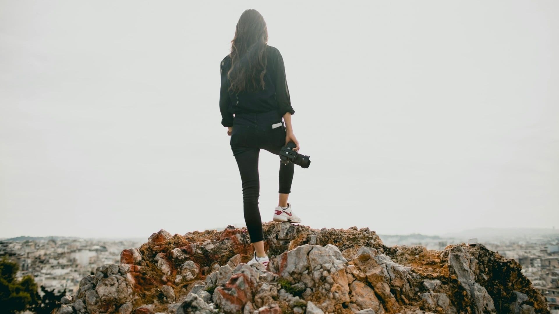 A woman in a black jacket, holding a camera and standing on rocky mountain overlooking a city – via Rachel Martin on Unsplash