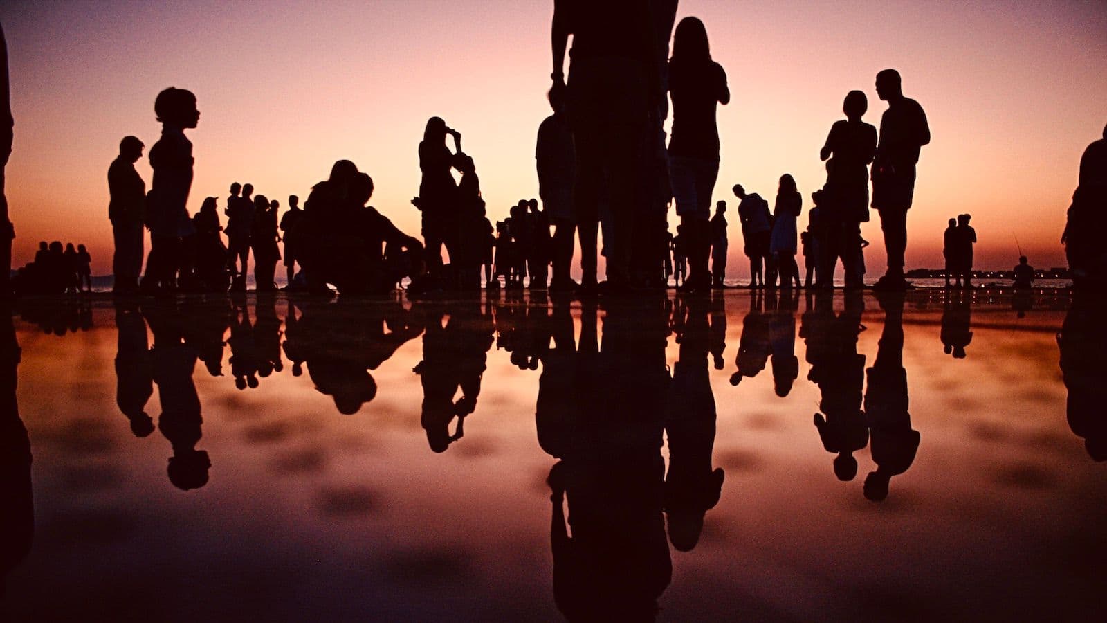 Picture (by Mario Purisic) shows the silhouettes of a large group of people standing and sitting on a beach at sunset.